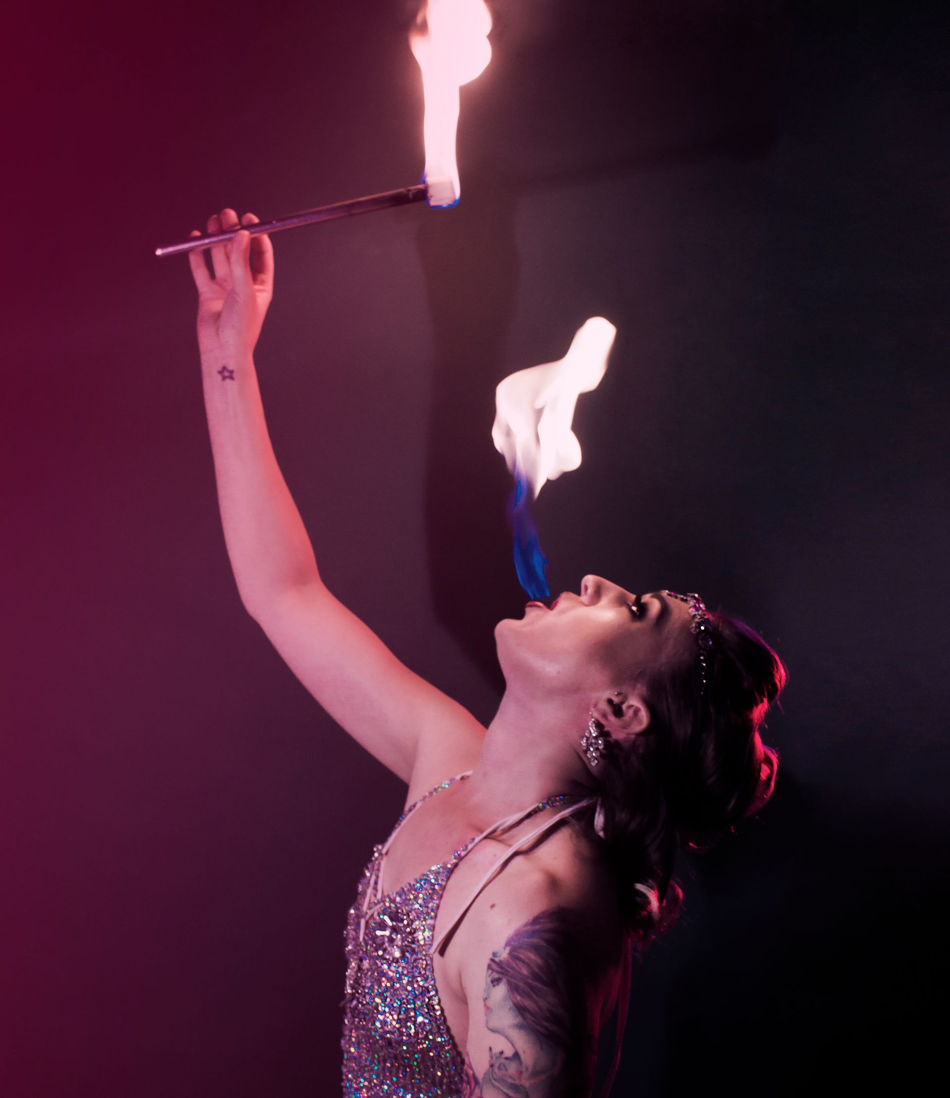 a posed colour photo of Mia Meow from the waist up with a dark pinkish background, she is holding a lit fire wand above her head and is performing a human candle with a large flame coming from her mouth by Snookie Mono Fauxtography
