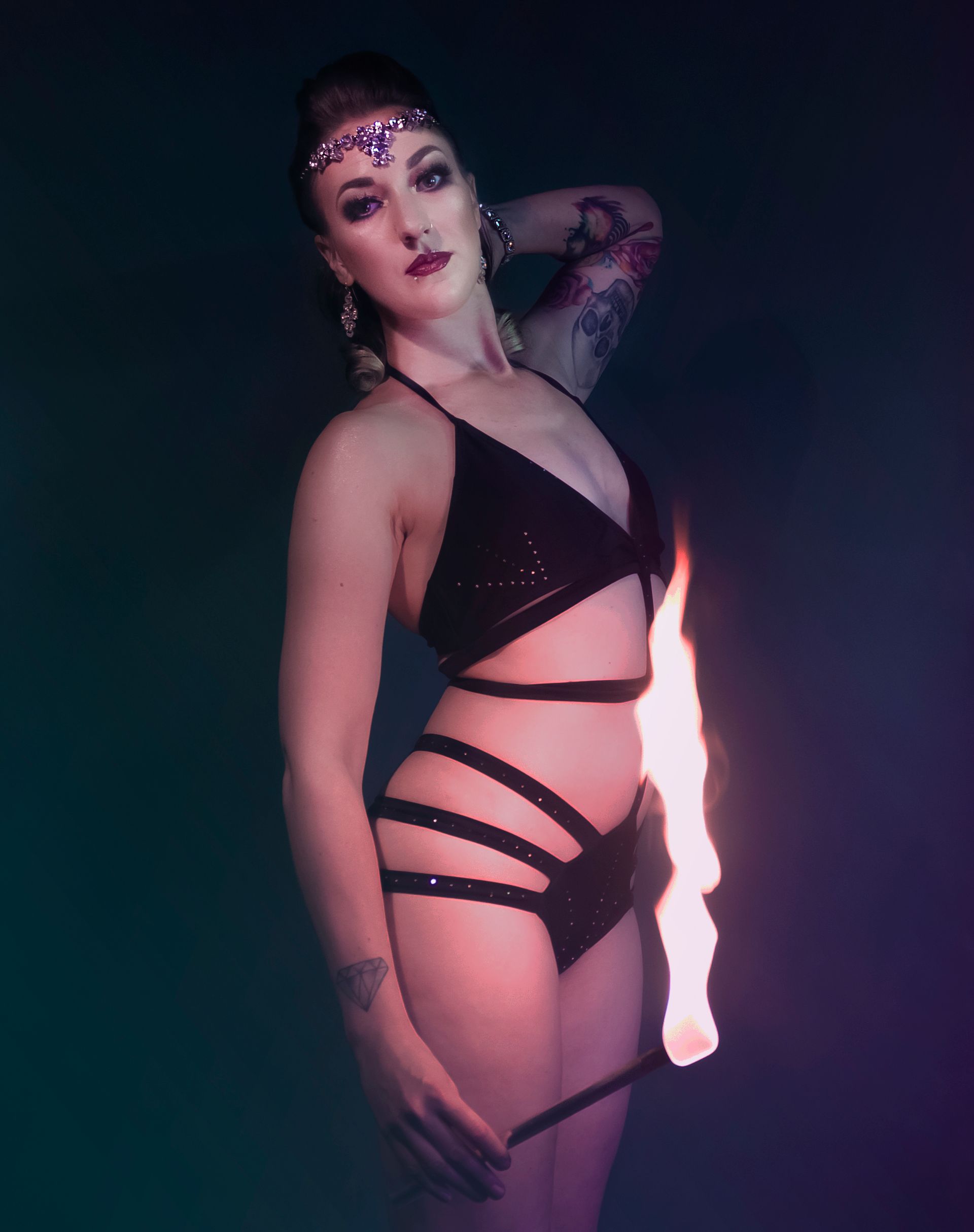 a colour image of Mia Meow in a posed photo wearing a black cage style costume one hand is behind her head the other is by her side holding a lit fire wand with the flame coming up in front of her by Snookie Mono Fauxtography