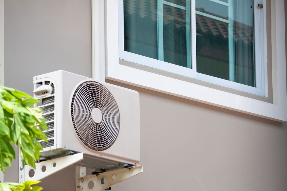 White air conditioner unit mounted on a gray wall next to a window; green plant in the foreground.