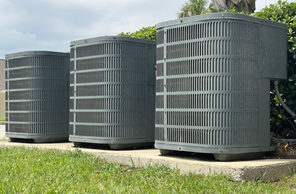 Three gray air conditioning units sitting on a concrete pad near green grass and bushes.