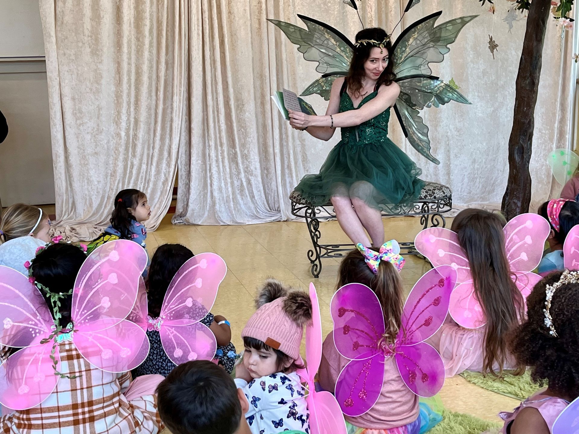 A woman dressed as a fairy is sitting on a bench talking to a group of children.