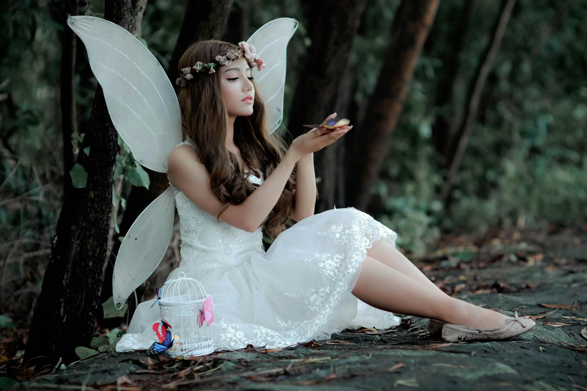 A woman dressed as a fairy is sitting under a tree in the woods.