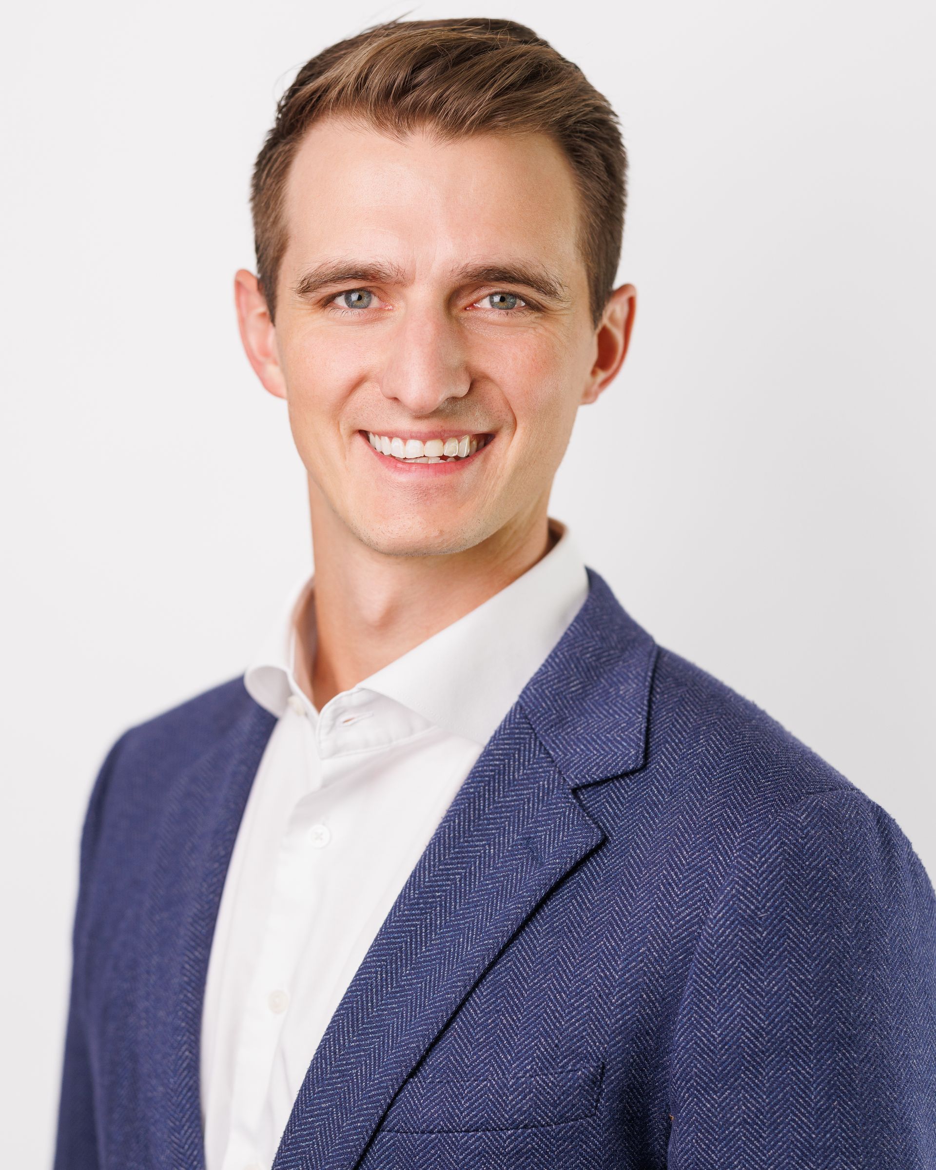 Man smiling, wearing a white shirt and blue blazer, against a white background.