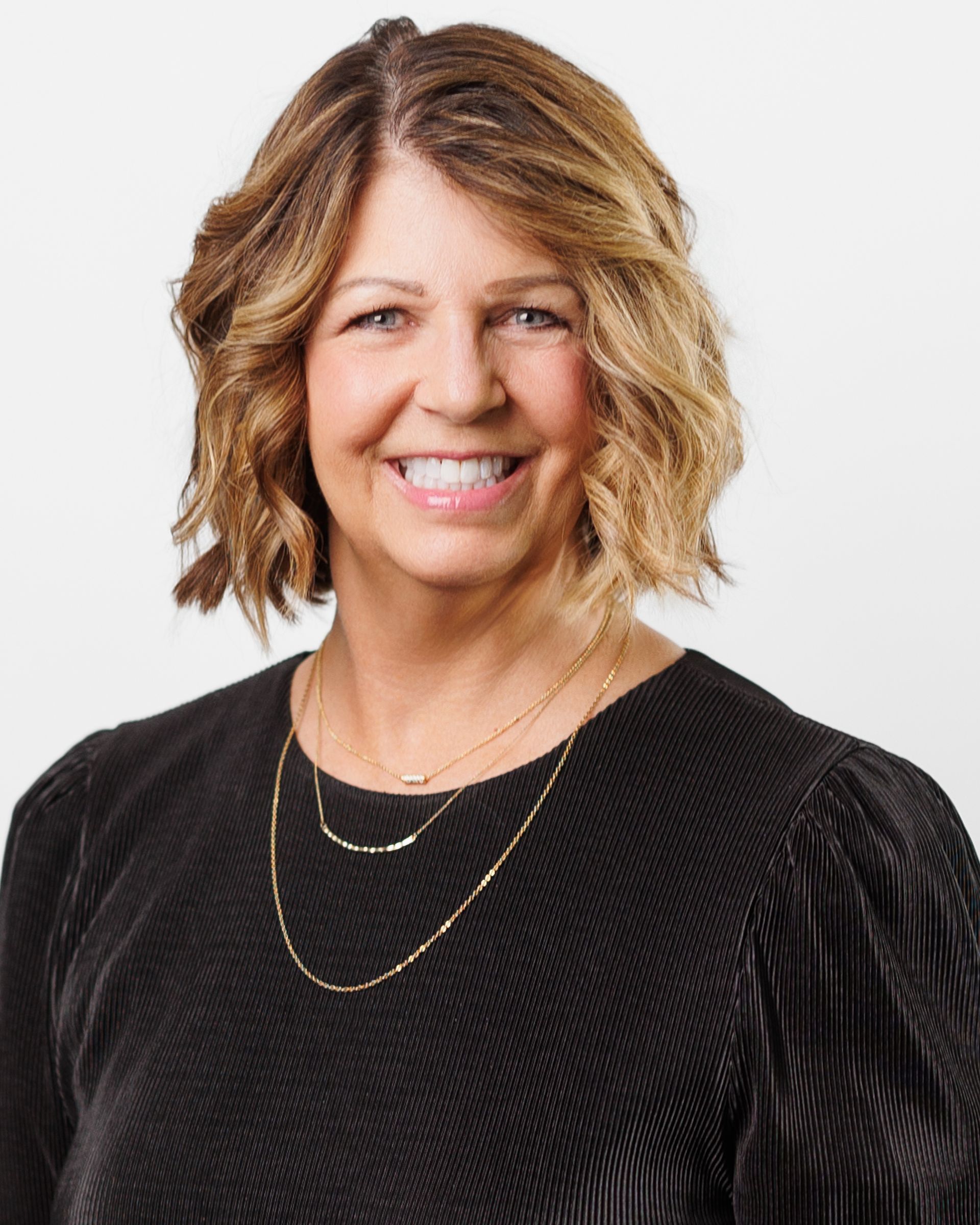 Woman with shoulder-length blonde hair, smiling at the camera, wearing a black top and gold necklace.