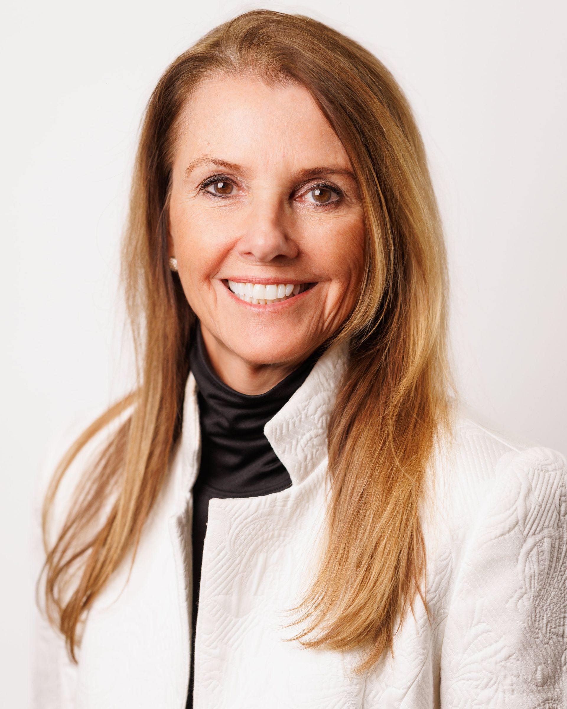 Woman with long, light brown hair smiles, wearing a black turtleneck and white blazer, against a white background.
