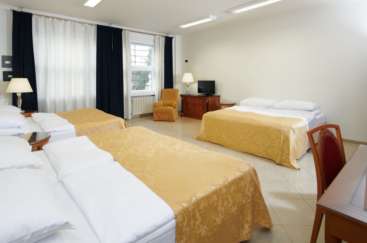 A hotel room with three beds, a desk, a TV, and an armchair. The room is white with gold and dark blue accents.