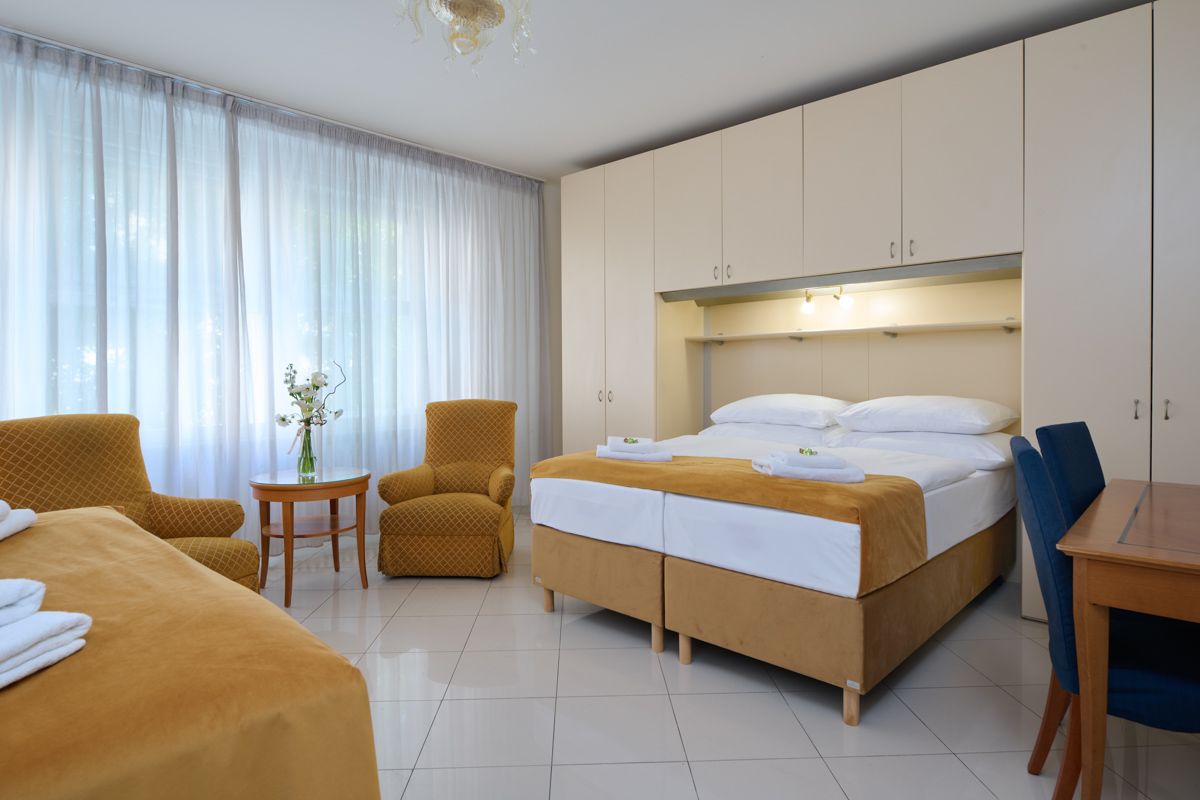Hotel room with two beds, two armchairs, and a table with a vase of flowers. Light yellow and beige colors.