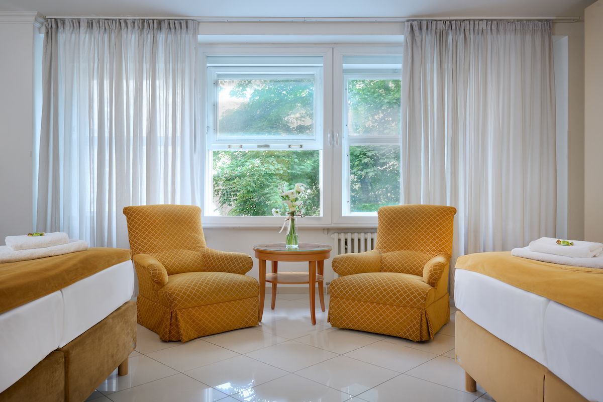Two twin beds, two yellow armchairs, and a table in a bright room with a large window and sheer white curtains.