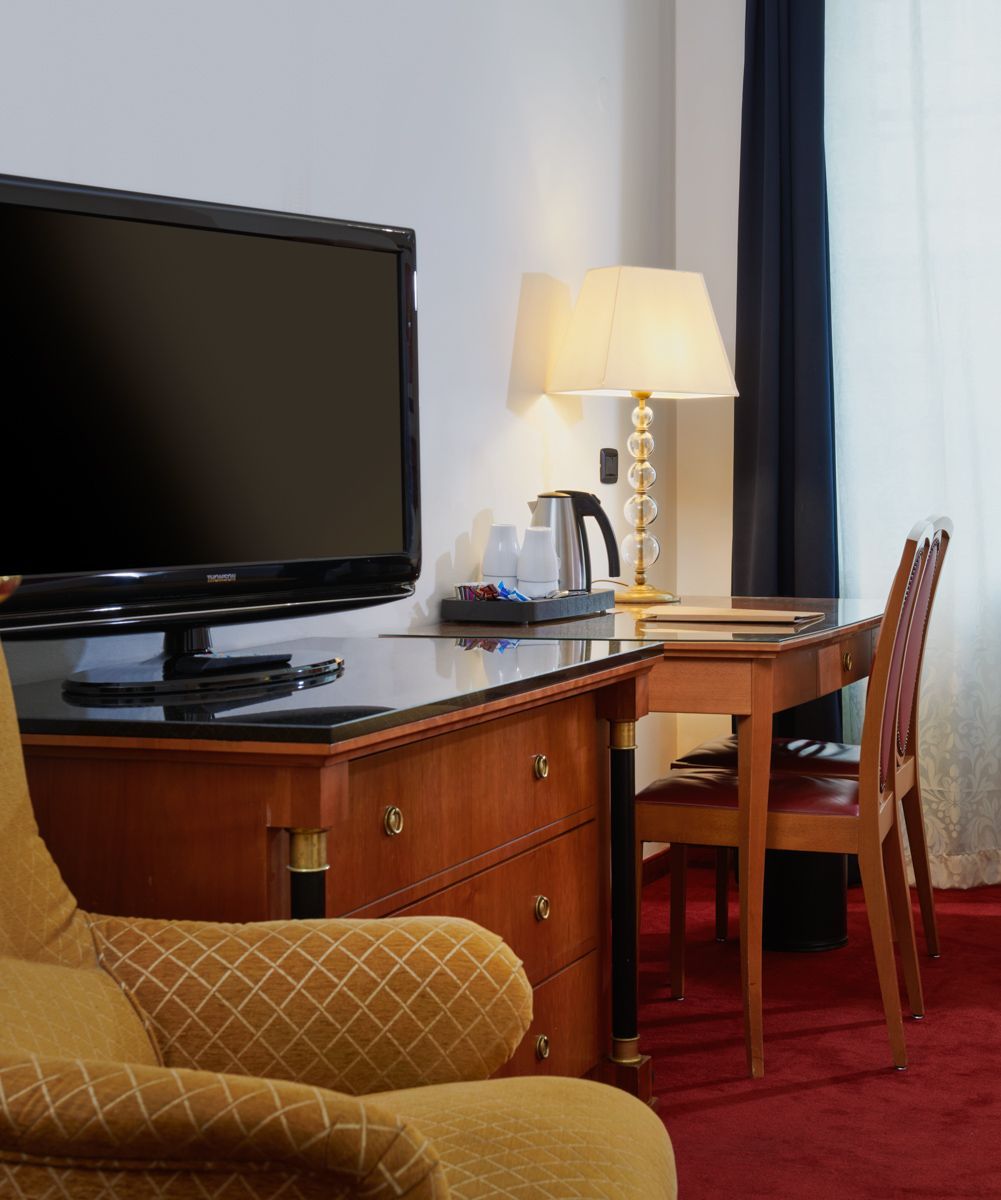 Hotel room corner with TV, desk, chair, lamp, and armchair on red carpet.