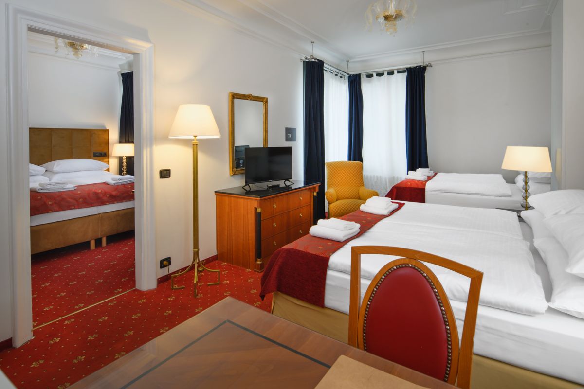 Hotel room with two beds, a desk, and a connecting room with another bed. Red carpet.
