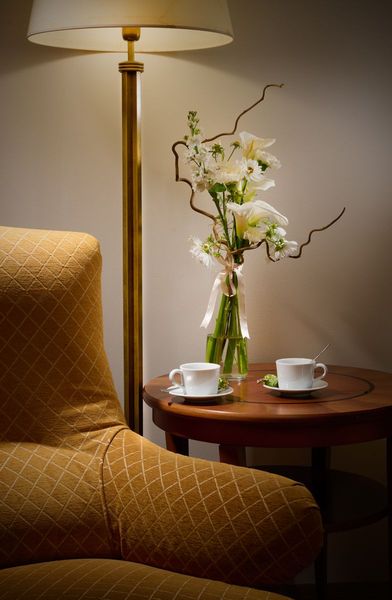 Cozy corner: upholstered chair, small round table with teacups, vase of white flowers, floor lamp.