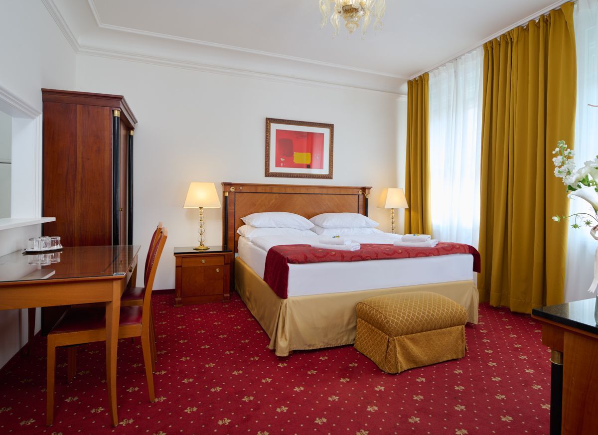Hotel bedroom with red carpet, double bed, desk, and golden curtains.