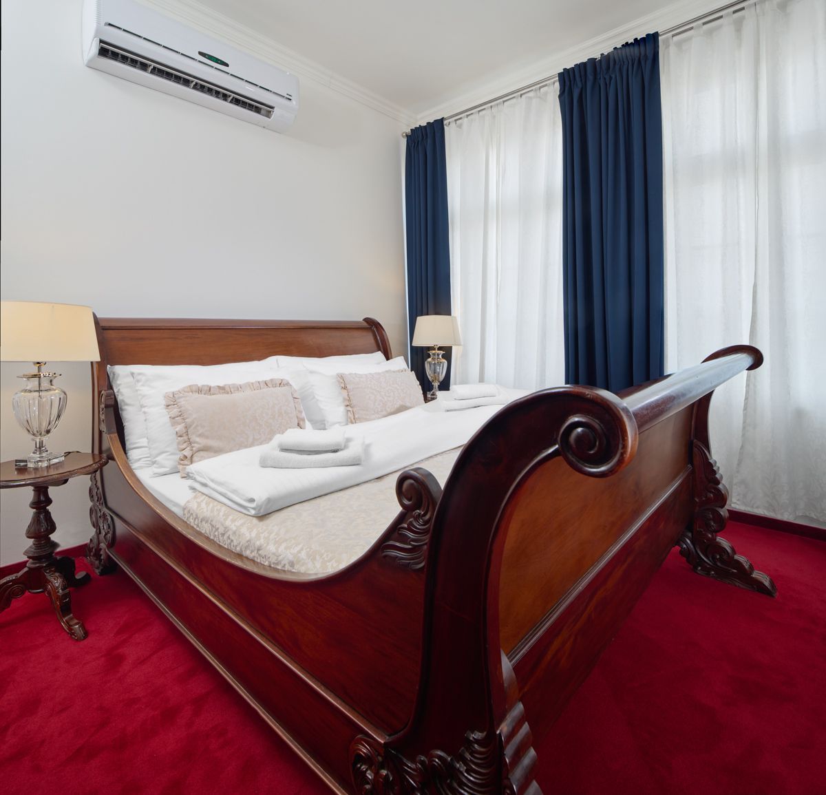 Bedroom with ornate wooden bed, red carpet, white walls, and blue curtains.