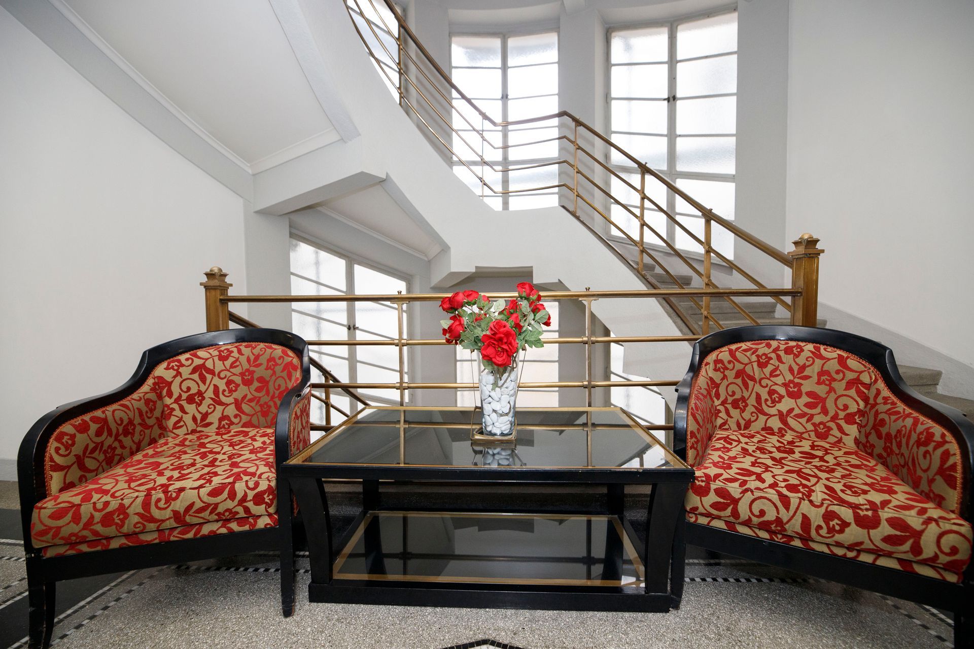 Two patterned chairs and table with flowers, under a staircase in a building's interior.