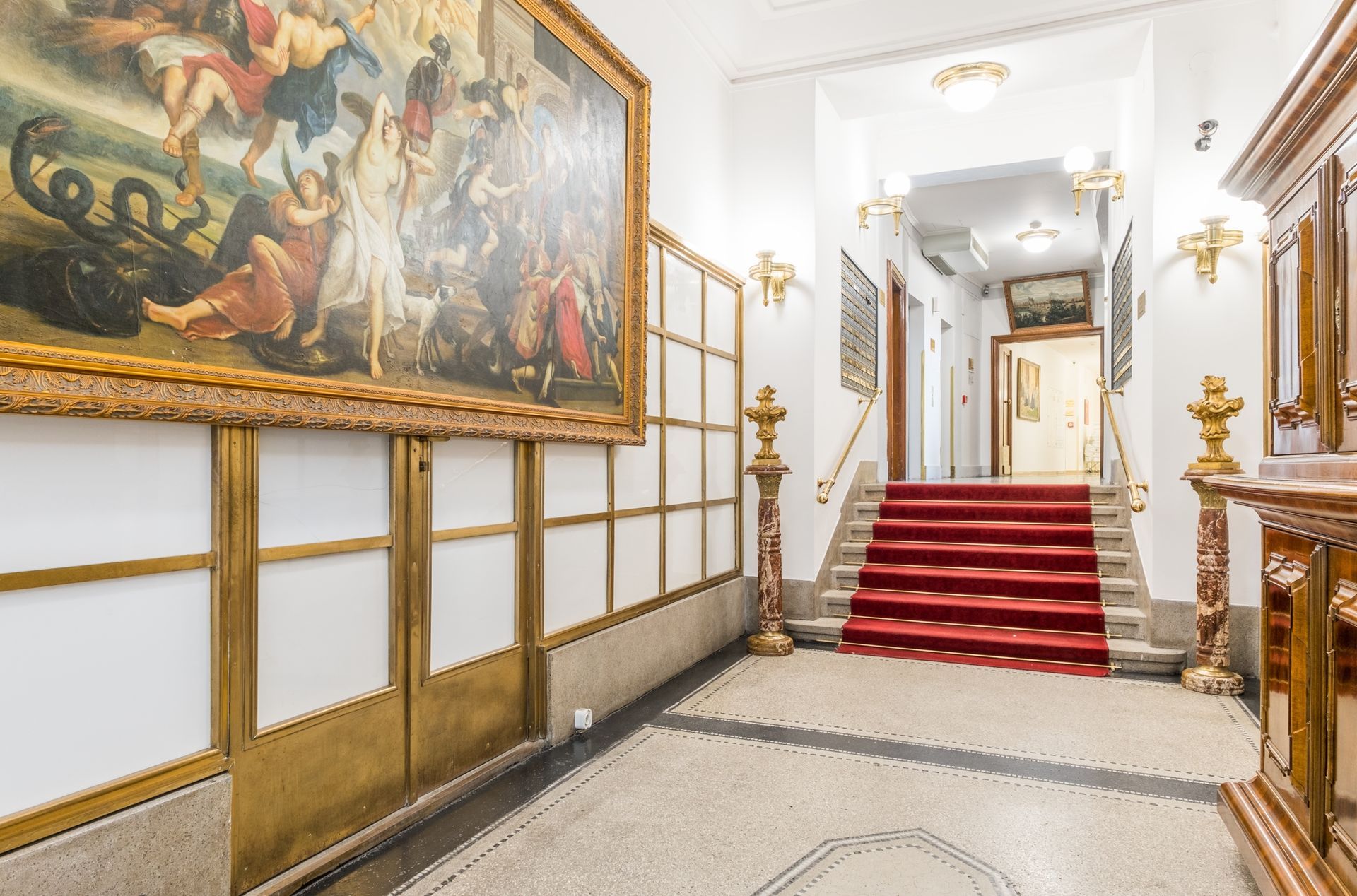 Ornate hallway with large painting, red-carpeted stairs, marble accents, and gold trim.