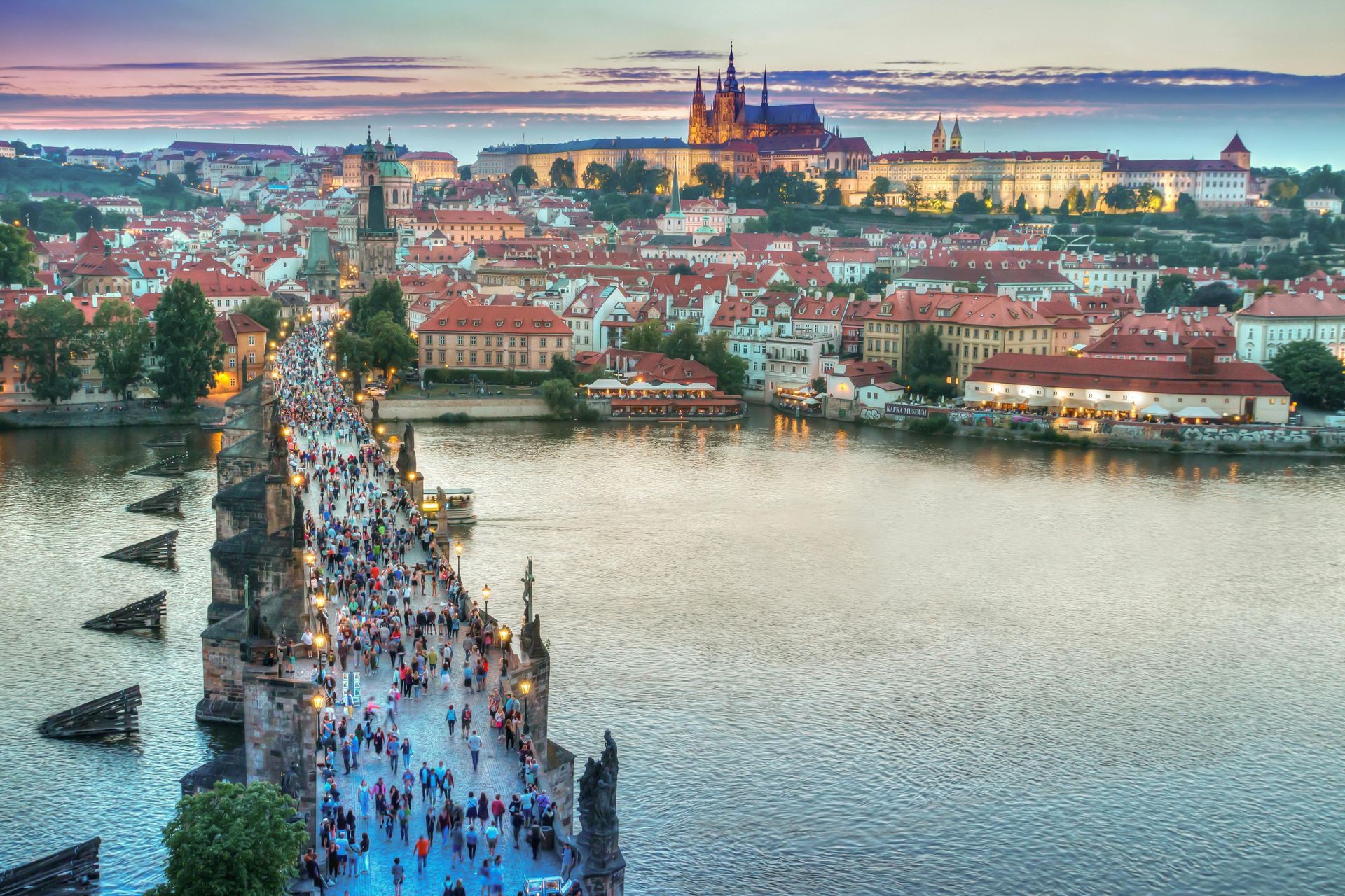 Prague cityscape at dusk, Charles Bridge with crowds, castle atop a hill, calm river.