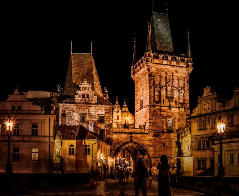 Night view of Prague's Old Town Bridge Tower, lit by warm lights. People walk along the bridge towards the tower.
