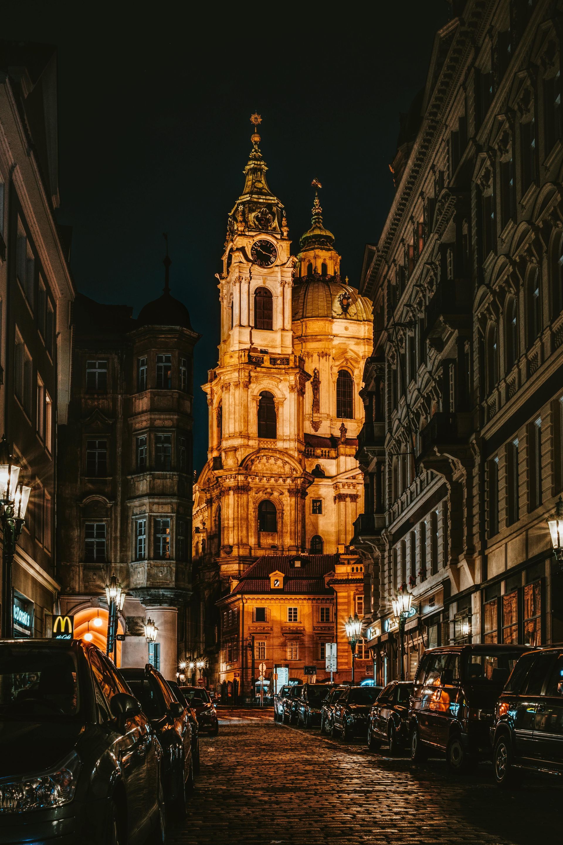 Lit-up church at night in Prague, Czech Republic, between buildings; cobblestone street, parked cars.