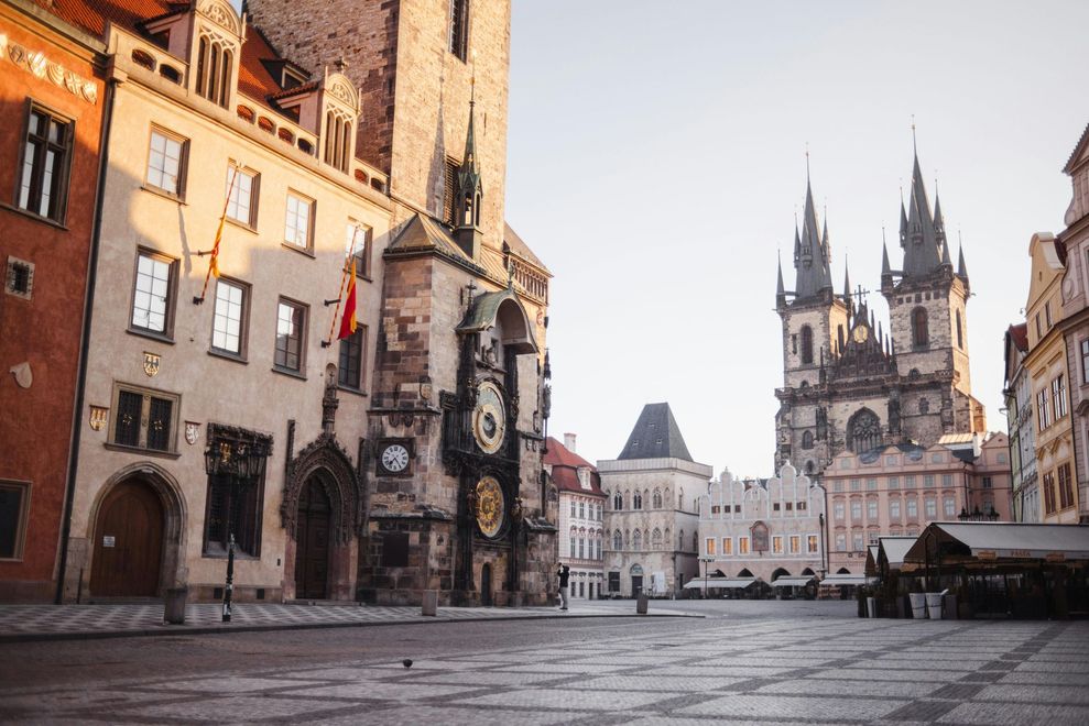 Buildings in a city square, featuring a clock tower and a church with spires. Warm sunlight.