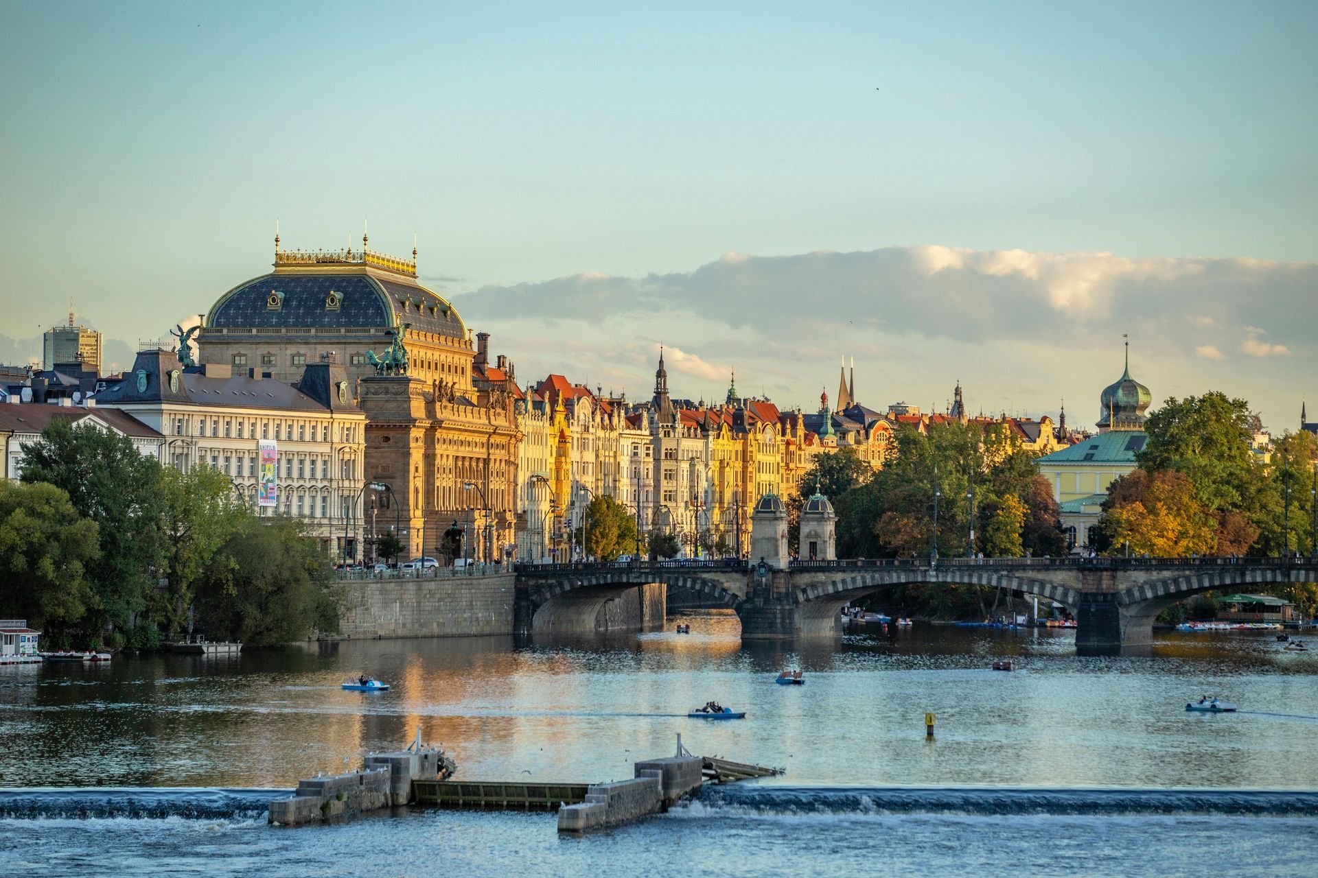 Prague cityscape at sunset, featuring a river with boats, a bridge, and buildings with golden hues.
