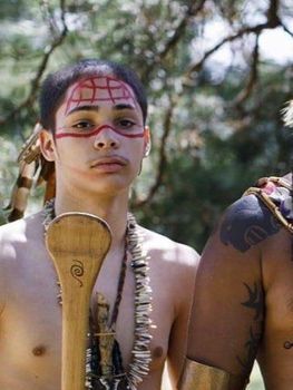 Miguel Wanderingturtle Garate dressed in traditional clothing and facepaint, holding a paddle. 