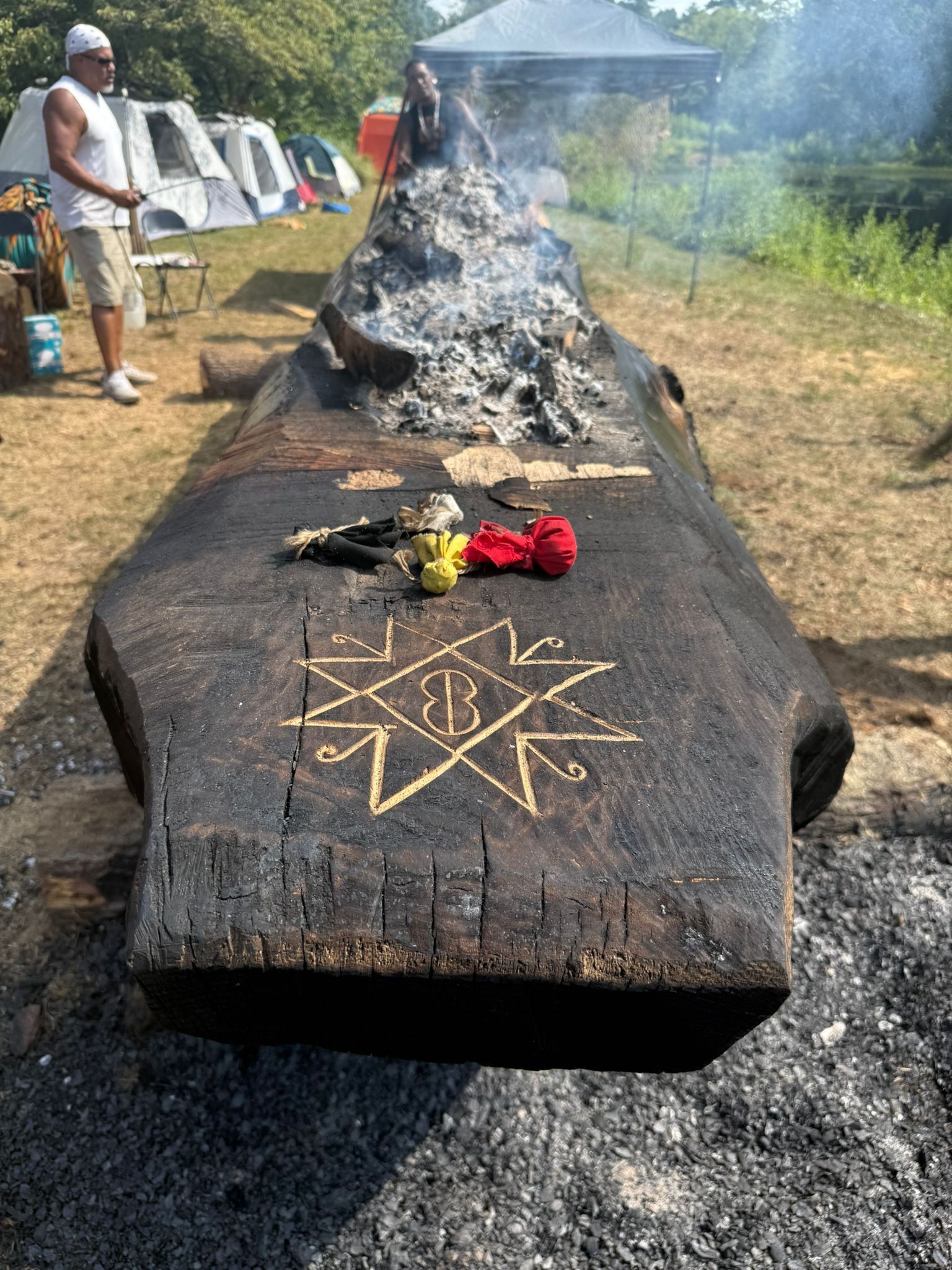 Long, charred log with a carved star symbol and offerings. People, tents, and smoke in the background.