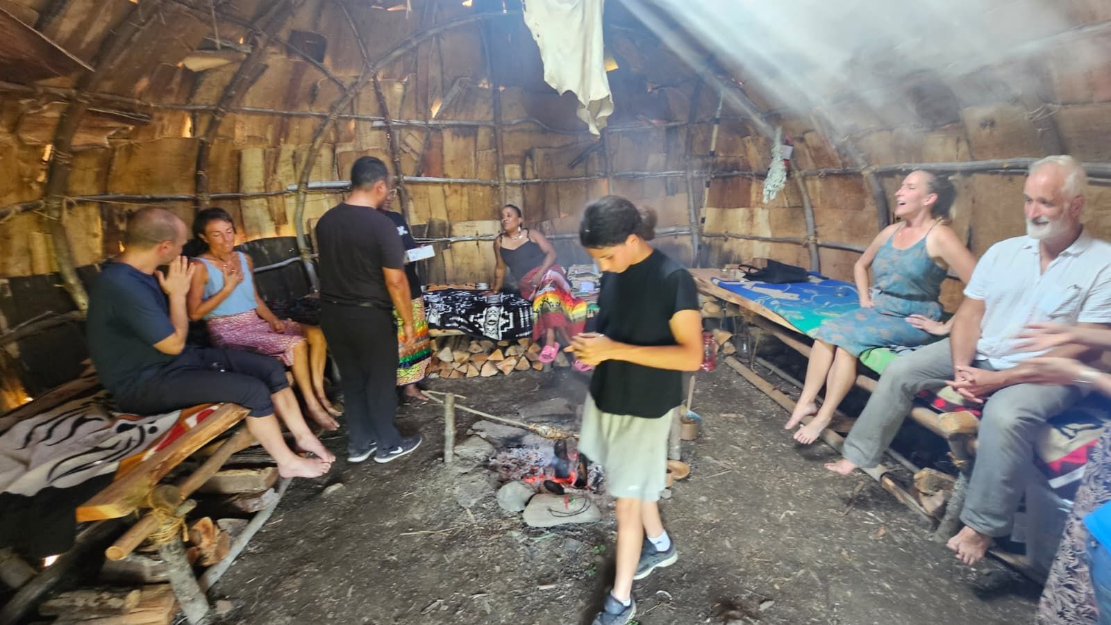 People gather inside a wooden structure, around a central fire. Smoke fills the air.