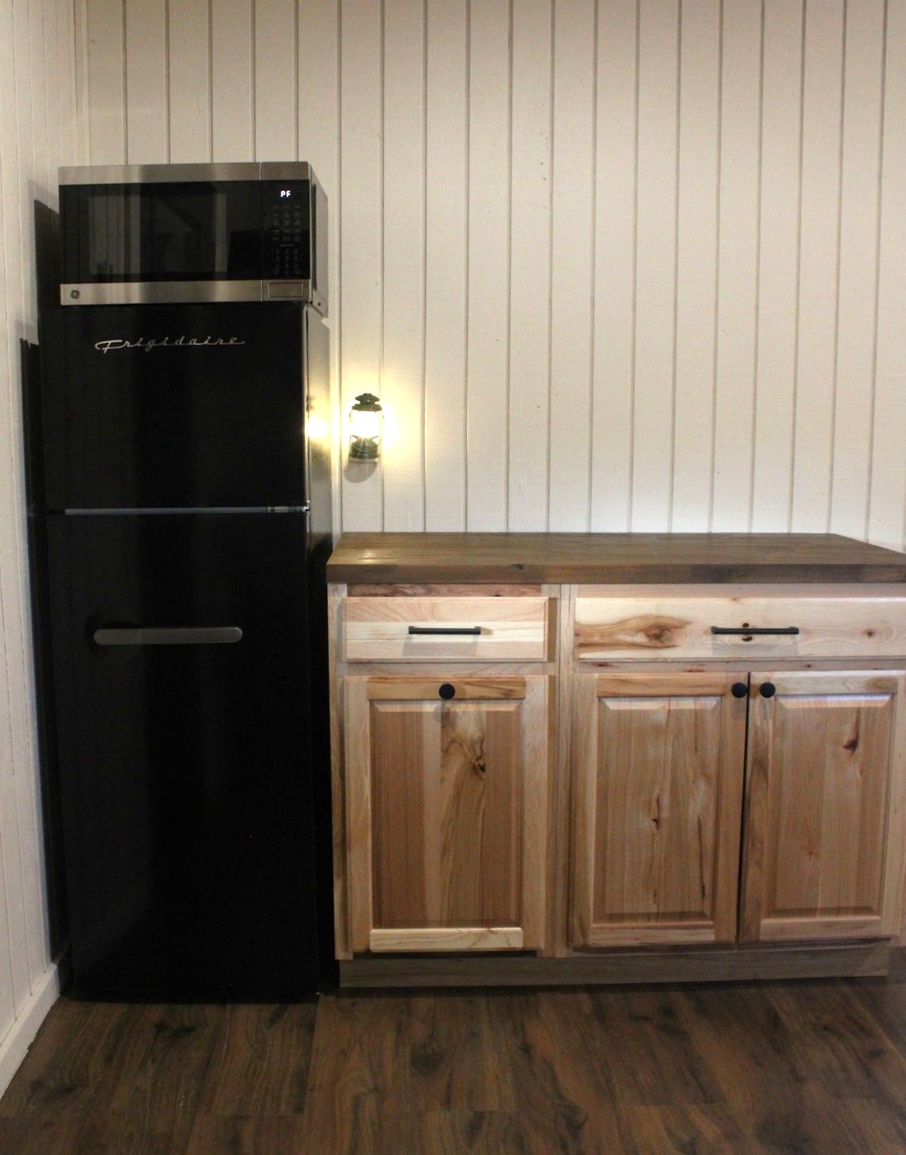 Black refrigerator beside wood kitchen cabinets with a lit wall sconce on a white paneled wall
