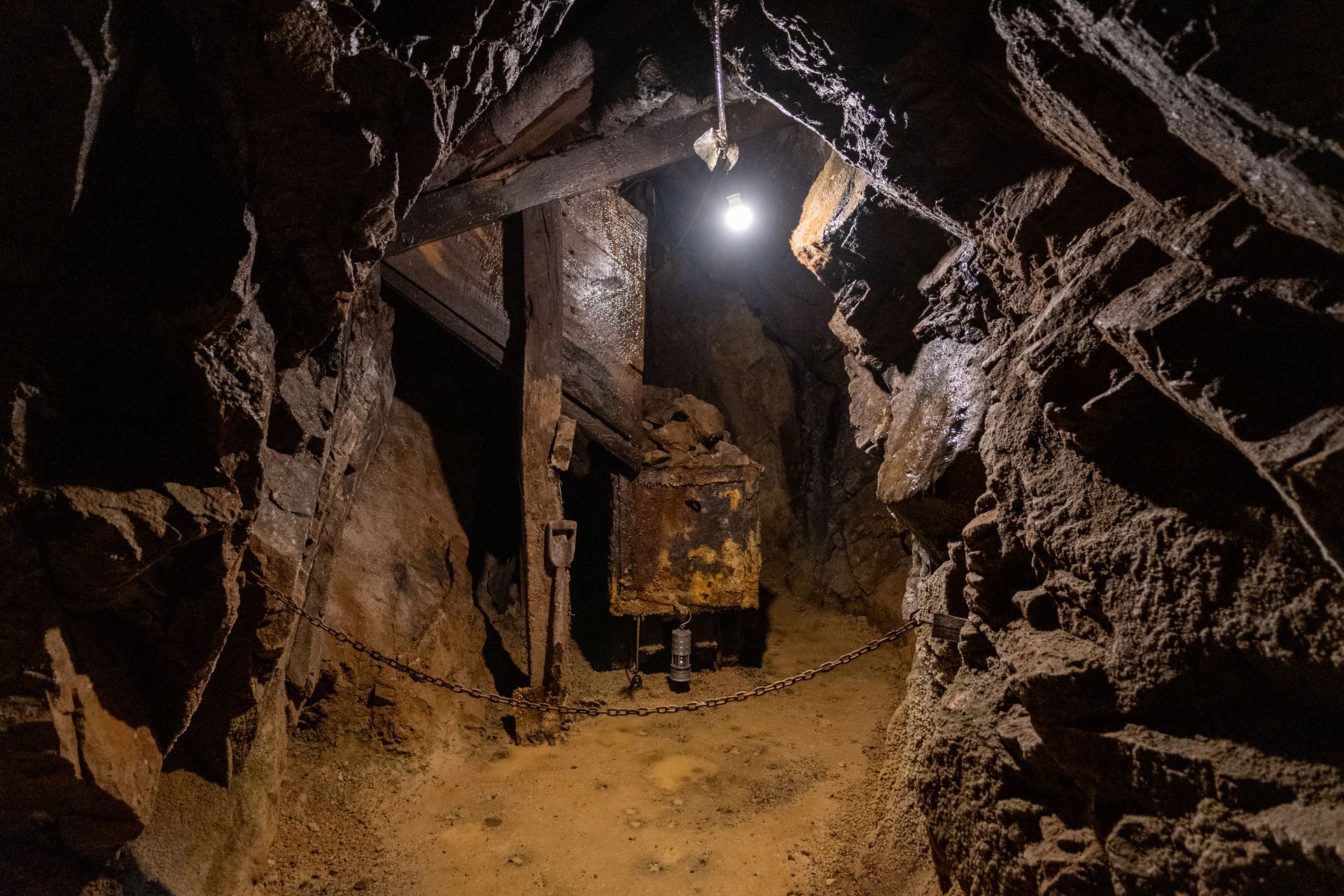 Dark, narrow mine shaft interior with wooden supports and a rusted mechanical device. A lightbulb illuminates the scene.
