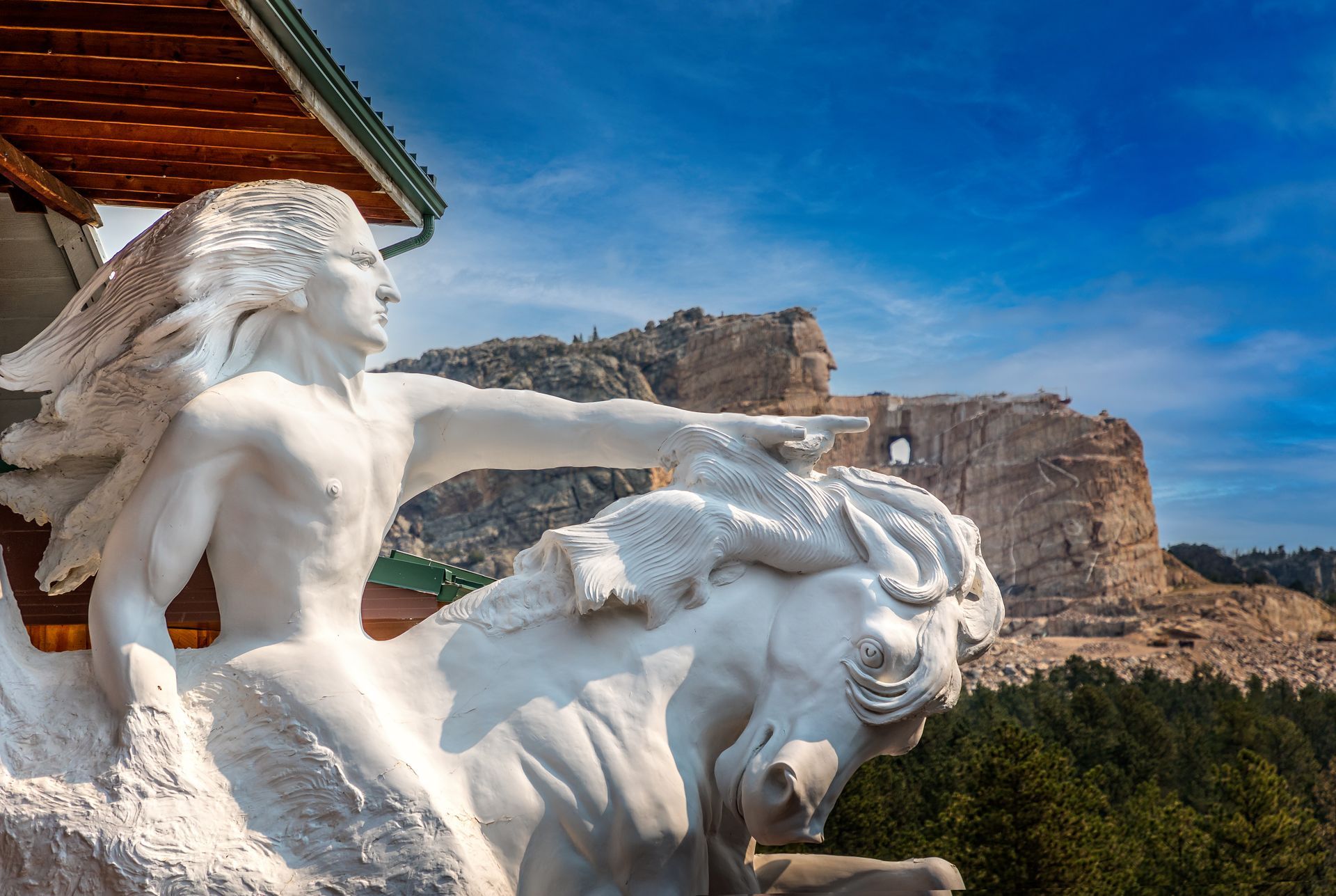 Sculpture of a Native American on horseback, pointing, with the unfinished Crazy Horse Memorial in the background.