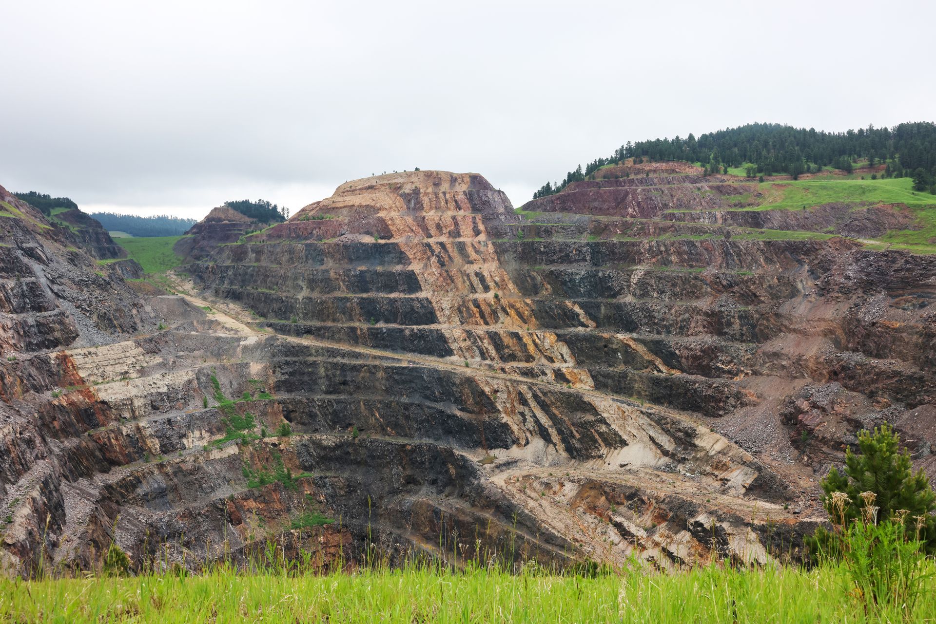 Open-pit mine with tiered rock and earth. Green grass in foreground, cloudy sky.