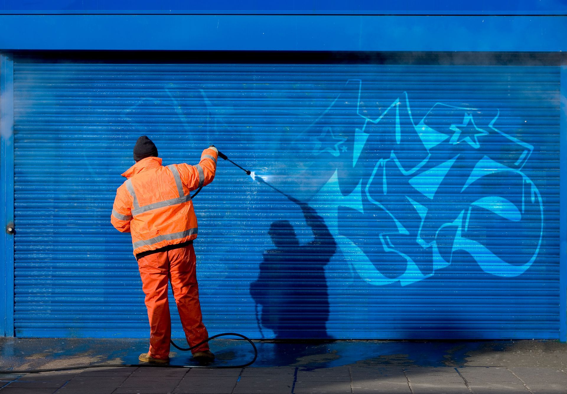 A man is cleaning graffiti on a blue garage door with a high pressure washer.