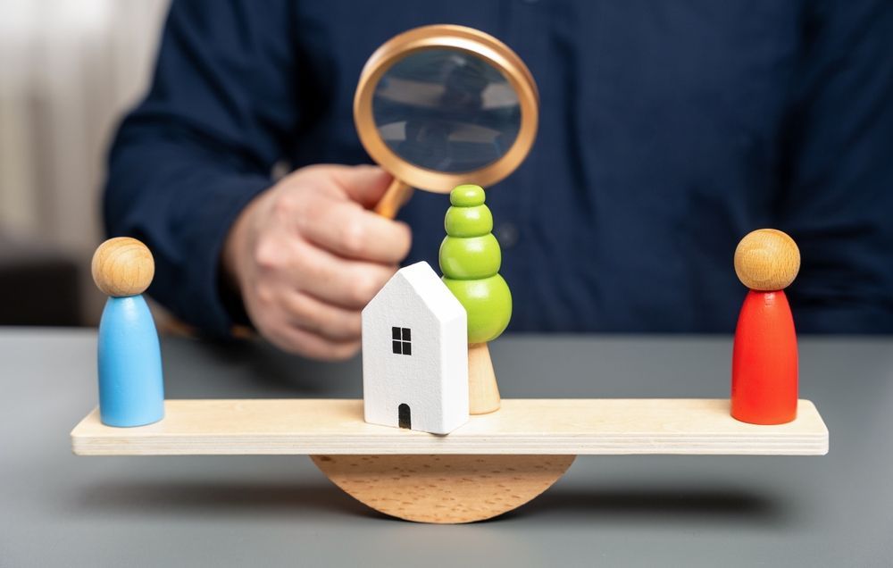 A person is holding a magnifying glass over a wooden seesaw with a house and two people on it.