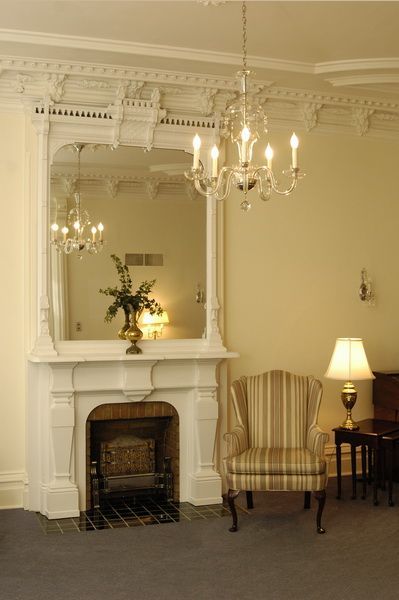 An elegant room with fireplace, mirror, chandelier, armchair, and lamp. Cream walls, ornate molding.