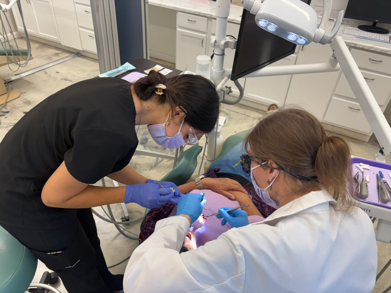 Two dental professionals in a clinic examine a patient, using tools and wearing masks and gloves.