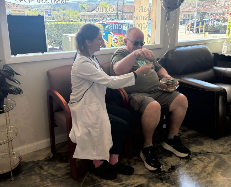 A person in a doctor's coat hands a small item to a seated person, near a window with balloons.