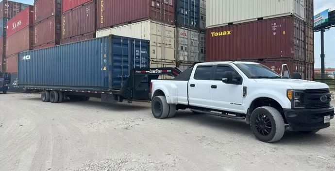 White pickup truck pulling a large blue shipping container on a trailer; parked near stacks of shipping containers.