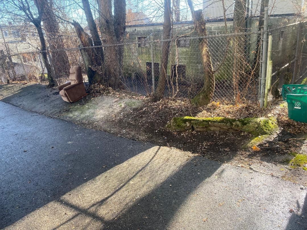 A green trash can is sitting on the side of a road next to a chain link fence.