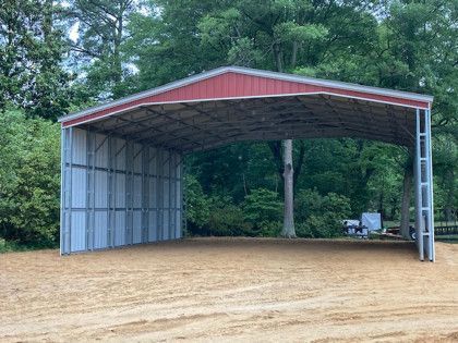 Metal carport with a red roof, located outdoors on a gravel lot. Trees in background.