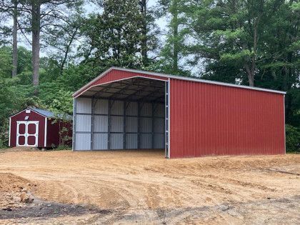 Red metal barn with open entrance next to a small wooden shed, both on a dirt lot.
