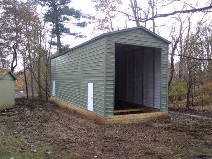 Green metal shed with large opening, small white doors, in a wooded area.