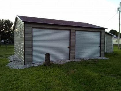 Tan metal two-car garage with two roll-up doors, a brown roof, and a gravel base, set on green grass.
