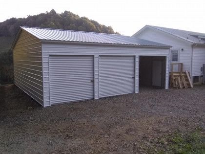 Metal garage with two doors and a side door, situated next to a house on gravel.