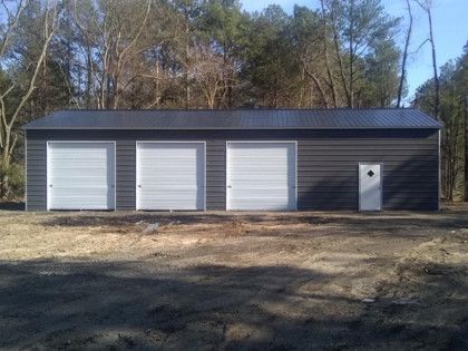 Three-bay, gray metal garage with white doors, black roof, and small side door, set in a wooded area.