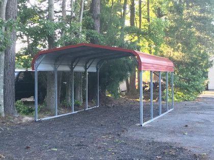 Metal carport with red trim, protecting a dark-colored vehicle on gravel driveway, surrounded by trees.