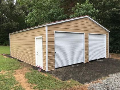 Tan metal two-car garage with white doors, side entry, set on asphalt, surrounded by greenery.