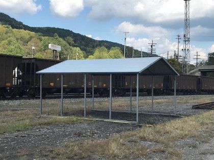 A metal-framed shelter with a silver roof, beside a train on a gravel lot, with a mountain backdrop.