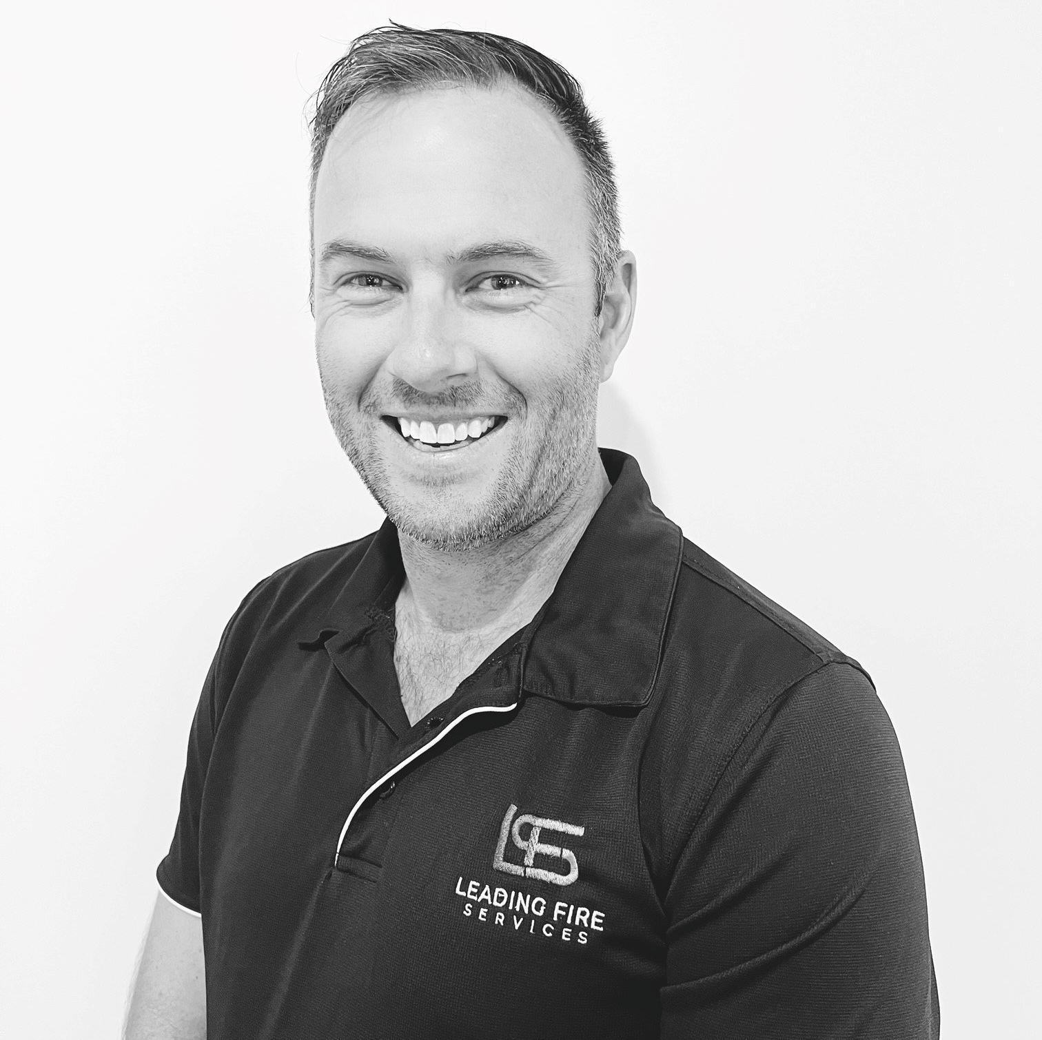 A man wearing a black shirt with the letter s on it is smiling in a black and white photo.