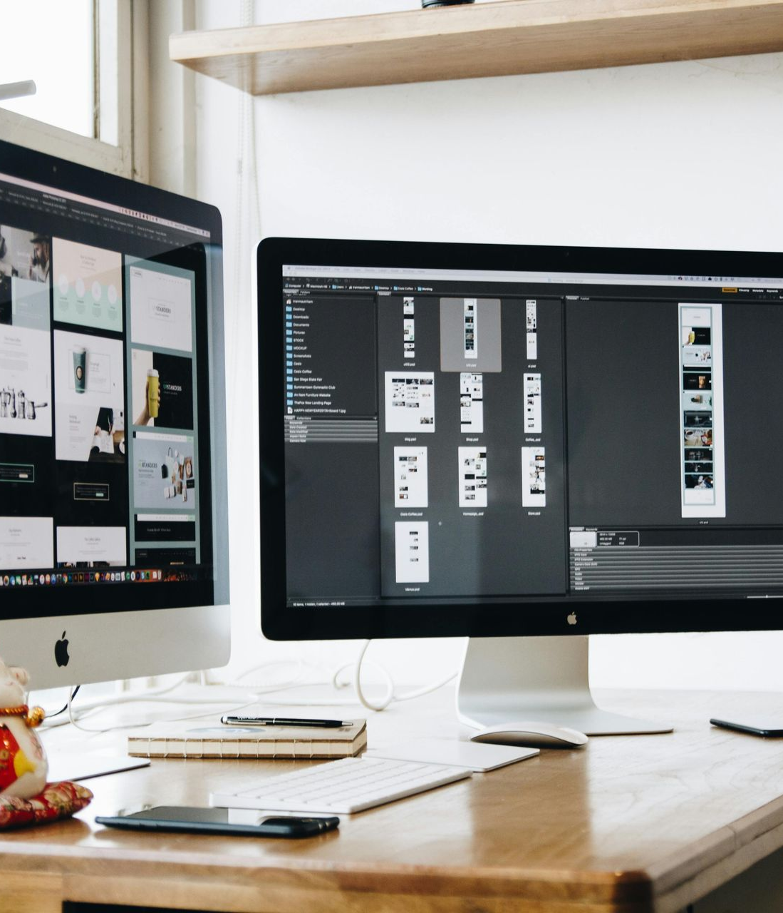 Two computer monitors on a wooden desk display graphic design work. A smartphone, notebook, and a small cat.
