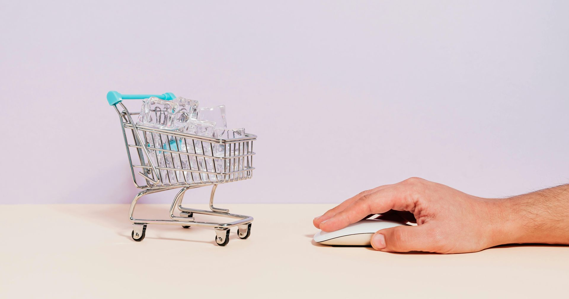Hand using a mouse next to a miniature shopping cart filled with items, suggesting online shopping.